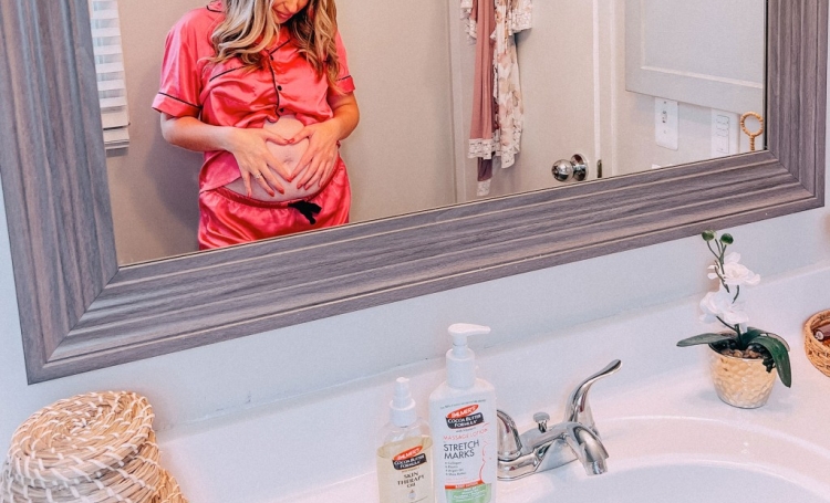 Woman standing at bathroom mirror with Palmer's Massage Lotion and Skin Therapy Oil looking at her pregnant belly during her second trimester self care routine